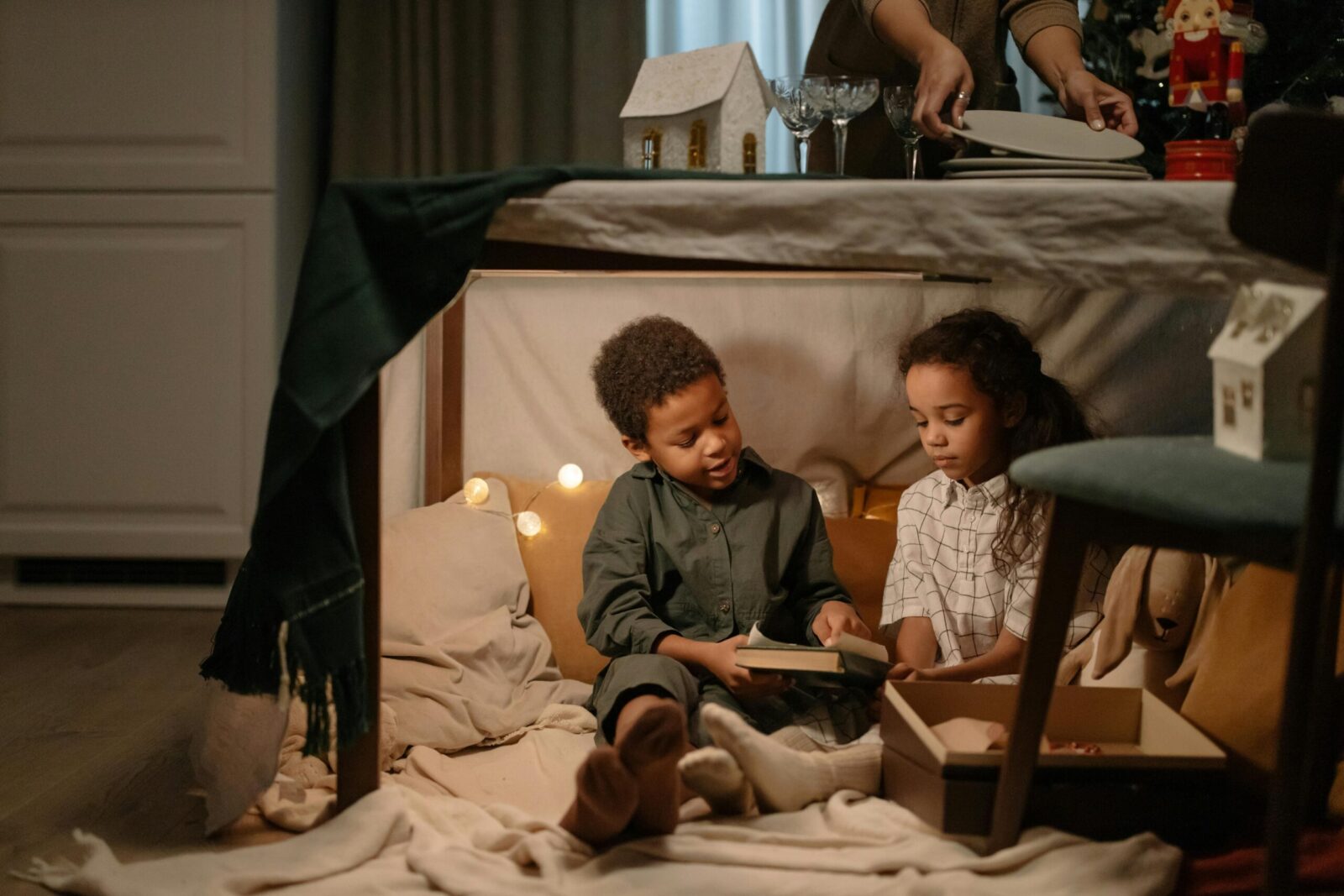 Two children enjoy reading together inside a cozy fort under a table with festive decorations.