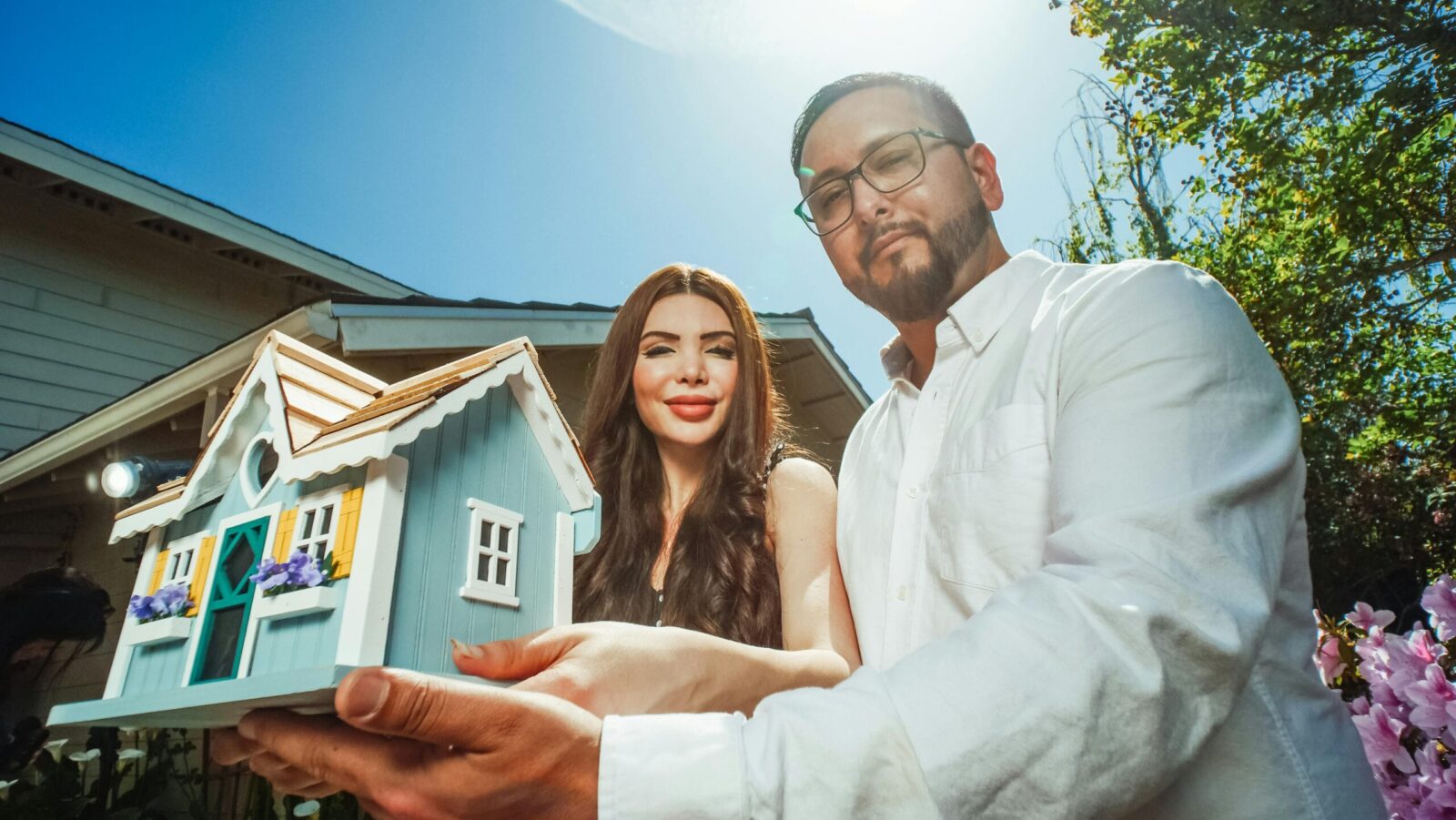 A happy couple holding a miniature house model outdoors, symbolizing new home ownership.